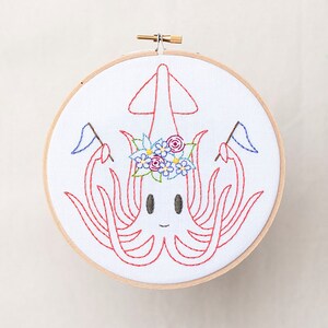 May include: Embroidered hoop art featuring a red squid with a flower crown and holding two blue flags. The squid has a smiling face and is surrounded by a white background.