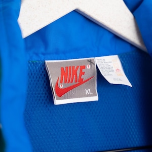 May include: Close-up of a blue jacket with a mesh lining, hanging on a white hanger. The Nike logo is visible on a tag, with the word "NIKE" in red and the size "XL". The jacket is made of a smooth, shiny material.