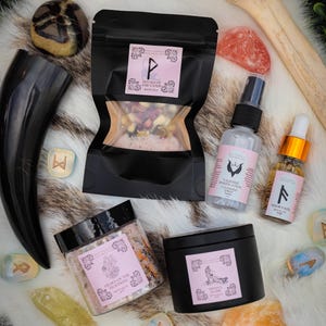 May include: An assortment of spiritual and self-care products, including a black horn, crystals, a black bag of bath tea, a spray bottle, a small bottle of oil, a jar of bath salts, and a black candle. The items are arranged on a white fur-like surface.