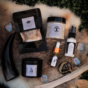 Puede incluir: Un surtido de productos espirituales y de bienestar, que incluyen una vela negra, una bolsa negra de t&eacute;, un tarro de sales de ba&ntilde;o y una botella de aceite. Los art&iacute;culos est&aacute;n dispuestos sobre una superficie similar a la piel con runas y un hueso.