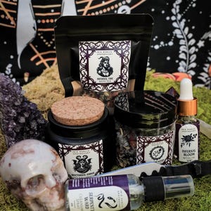 May include: A collection of witchy bath and body products, including a bath tea, a soy candle, a ritual oil, and a cleansing spray. The products are packaged in black jars and bottles with white labels featuring gothic illustrations and text. The text on the labels includes "Infernal Tide Bath Tea", "Nocturnal Ember Soy Candle", "Infernal Bloom Ritual Oil", and "Serpents Ward Cleansing Spray".
