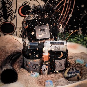 Puede incluir: Una bolsa de malla negra llena de varios art&iacute;culos de boticario, incluyendo frascos, botellas y un peque&ntilde;o vial. La bolsa est&aacute; decorada con estrellas doradas y lunas crecientes. Los art&iacute;culos est&aacute;n dispuestos sobre una superficie similar a la piel.