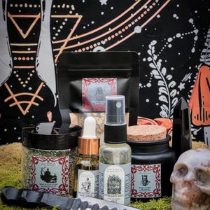 May include: An assortment of apothecary products, including jars, bottles, and a black bag, arranged on a bed of green moss. A decorative knife and a skull are also present. The products have labels with gothic-style illustrations.