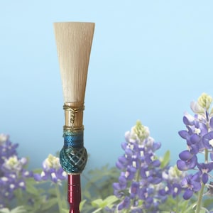 Puede incluir: Primer plano de la caña de un instrumento de lengüeta, con una cabeza de caña de color beige claro en forma de trapecio. La caña está adornada con alambre dorado, hilo azul y verde y una base roja. El fondo es azul suave con flores moradas.