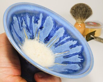 May include: A blue ceramic shaving bowl with a white, foamy substance inside. The bowl has a unique design with blue and white patterns. A shaving brush, a razor, and a shaving soap are in the background.