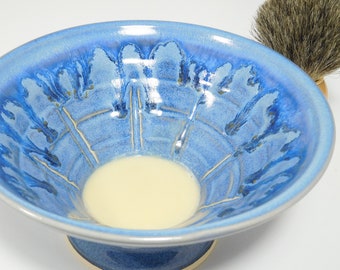 May include: A blue ceramic shaving bowl with a light cream-colored interior. The bowl has a pedestal base and is decorated with a floral pattern in shades of blue. A shaving brush with a wooden handle and dark bristles is visible.