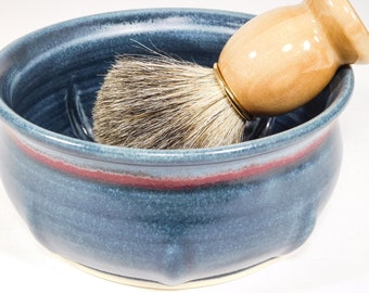 May include: A blue ceramic shaving bowl with a red stripe and a shaving brush. The bowl has a textured surface and a curved shape. The shaving brush has a wooden handle and a brush head made of bristles.
