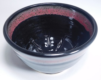 May include: A handmade ceramic bowl with a glossy black interior and a red and black rim. The bowl has a textured surface and a wide, open shape, suitable for serving or display.