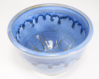 May include: A handmade ceramic bowl with a blue glaze. The bowl has a round shape with a slightly flared rim. The interior features a starburst design with gold accents. The rim has a dark blue drip pattern.