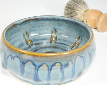 May include: A handmade ceramic shaving bowl in shades of blue and brown. The bowl has a textured surface with a decorative rim and interior accents. A shaving brush with a wooden handle and natural bristles rests beside the bowl.