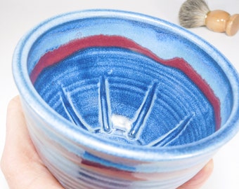 May include: A ceramic shaving bowl featuring a vibrant blue glaze with deep red accents. The interior of the bowl is textured with raised ridges designed to assist in lathering, and a small wooden shaving brush is visible in the background.