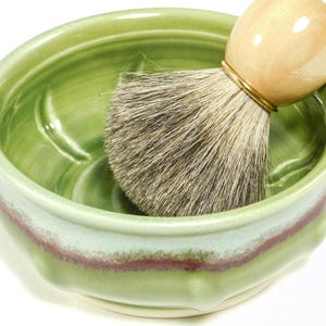 May include: A green ceramic shaving bowl with a light green and burgundy stripe. A shaving brush with a light wood handle and gray bristles rests inside the bowl. The bowl has a textured interior.