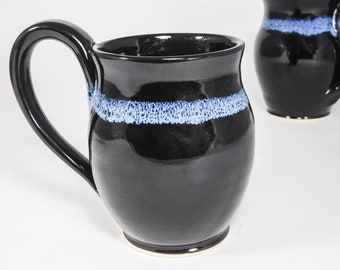 May include: Black ceramic mug with a large handle and a blue and white band around the middle. The mug has a glossy finish and a simple, elegant design. A second mug is visible in the background.