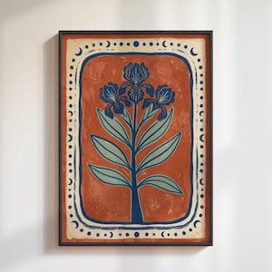May include: A framed rectangular wall art print featuring a central dark blue iris flower illustration with light green leaves on a textured terracotta background. The design is enclosed by a cream border decorated with dark blue moon phases and dots.