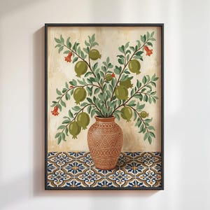 Op de afbeelding: Ingelijste kunstdruk met een stilleven van een vaas met groene granaatappels en bladeren. De bruine vaas heeft een geometrisch patroon. De achtergrond is lichtbeige en de basis heeft een blauw en wit tegelontwerp.