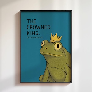 May include: Framed art print with a cartoon frog wearing a gold crown. The frog is green with large yellow eyes, set against a teal background. Text reads "THE CROWNED KING. OF THE WATER LILY."