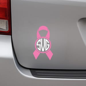 May include: A pink ribbon decal with a white circular monogram featuring the letters SVG. The decal is affixed to the rear of a silver vehicle. The ribbon design is a symbol of awareness.