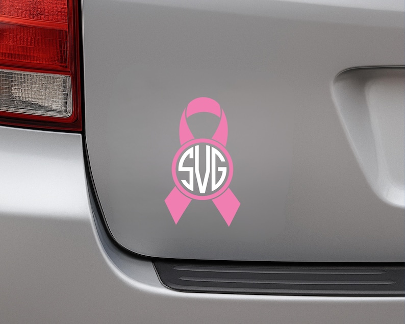 May include: A pink ribbon decal with a white circular monogram featuring the letters SVG. The decal is affixed to the rear of a silver vehicle. The ribbon design is a symbol of awareness.