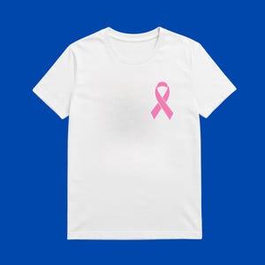 May include: A white t-shirt featuring a pink awareness ribbon on the left breast. The short-sleeved top is made of a soft material and is set against a blue background. The ribbon is a symbol of support.