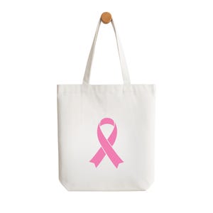 May include: A white tote bag featuring a pink awareness ribbon. The bag has two handles and is suspended against a white backdrop. The tote bag is made from a canvas-like material. The bag is designed for carrying items.