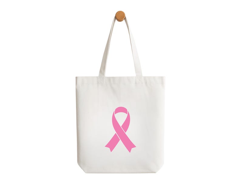May include: A white tote bag featuring a pink awareness ribbon. The bag has two handles and is suspended against a white backdrop. The tote bag is made from a canvas-like material. The bag is designed for carrying items.