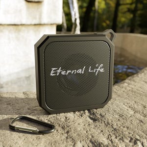 May include: A black, square-shaped portable speaker with the text "Eternal Life" in white script. The speaker has a perforated front and a loop for attaching a strap. A black carabiner clip rests below the speaker.