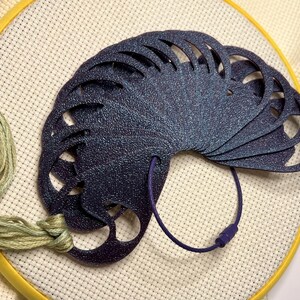 May include: A dark blue, fan-shaped needle minder with a textured surface, attached to a purple cord. The needle minder is on a white cross-stitch fabric stretched in a yellow embroidery hoop. Green embroidery floss is also visible.