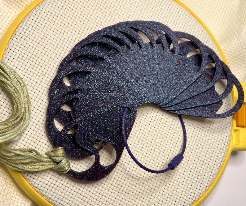 May include: A dark blue, fan-shaped needle minder with a textured surface, attached to a purple cord. The needle minder is on a white cross-stitch fabric stretched in a yellow embroidery hoop. Green embroidery floss is also visible.