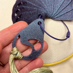 May include: A close-up of a blue and purple thread organizer with a fan-shaped design. The organizer is connected to a teardrop-shaped tag with a purple cord. Green and yellow embroidery floss is attached to the tag. A yellow embroidery hoop is in the background.