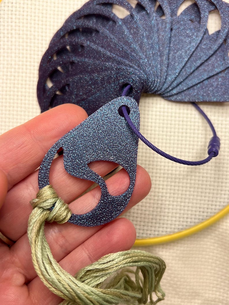 May include: A close-up of a blue and purple thread organizer with a fan-shaped design. The organizer is connected to a teardrop-shaped tag with a purple cord. Green and yellow embroidery floss is attached to the tag. A yellow embroidery hoop is in the background.