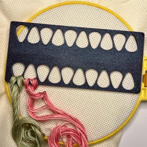Op de afbeelding: Een kruissteekproject in uitvoering, met een gele borduurring met witte stof. Een donkerblauwe draadorganizer met meerdere druppelvormige gaten is over de stof geplaatst. Groen en roze borduurgaren zijn zichtbaar.