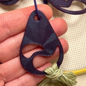 May include: A dark purple, teardrop-shaped pendant with a cut-out design, strung on a purple cord. The pendant is connected to a bundle of light green embroidery floss. The background is a cream-colored fabric.