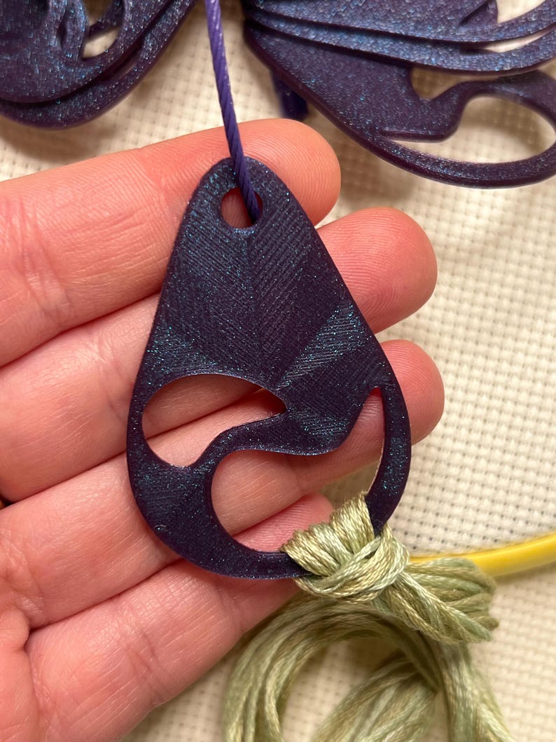 May include: A dark purple, teardrop-shaped pendant with a cut-out design, strung on a purple cord. The pendant is connected to a bundle of light green embroidery floss. The background is a cream-colored fabric.