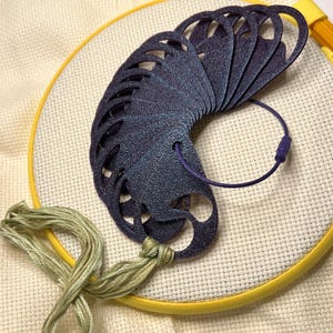 May include: A collection of dark blue, glittery, die-cut shapes, possibly for embroidery, arranged in a semi-circle. They are connected by a purple cord and sit on a white fabric stretched within a yellow embroidery hoop. Green and tan embroidery floss is also present.