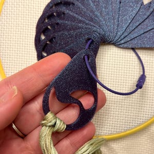May include: A dark blue, fan-shaped craft tool with a glittery finish, connected to a purple cord. The tool is held with a bundle of light green embroidery floss, and a yellow embroidery hoop is in the background. This tool is used for cross-stitch.