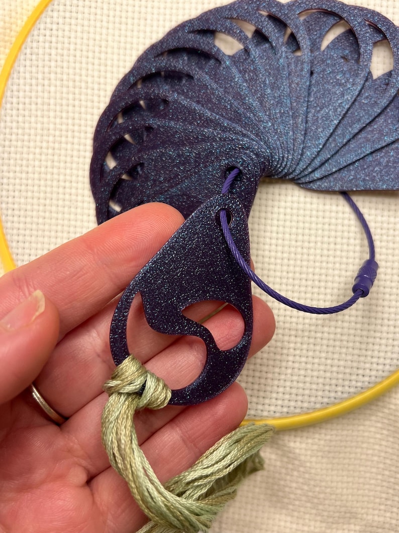 May include: A dark blue, fan-shaped craft tool with a glittery finish, connected to a purple cord. The tool is held with a bundle of light green embroidery floss, and a yellow embroidery hoop is in the background. This tool is used for cross-stitch.