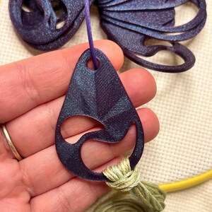 May include: A dark purple, teardrop-shaped pendant with a cut-out design, suspended by a purple string. The pendant is connected to a bundle of green thread. A larger, matching purple decorative element is visible in the background.