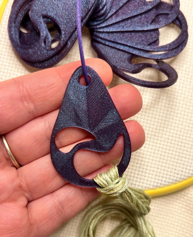 May include: A dark purple, teardrop-shaped pendant with a cut-out design, suspended by a purple string. The pendant is connected to a bundle of green thread. A larger, matching purple decorative element is visible in the background.