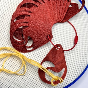 Op de afbeelding: Een stapel rode, lasergesneden leren stukken met decoratieve uitsparingen, verbonden door een rood koord. Een gele draad is door een rode leren hanger geregen. De items liggen op een witte stof met een blauwe rand.