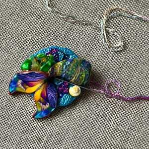 May include: A colorful butterfly-shaped charm with a blue, purple, yellow, and green design. The charm is attached to a blue and green background with a small white seashell.