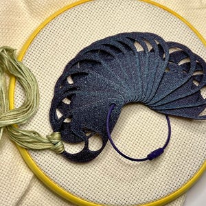 May include: A dark blue, fan-shaped needlework thread organizer with cut-out designs, held together by a purple cord and clasp. The organizer rests on a yellow embroidery hoop with cream-colored fabric and green thread.