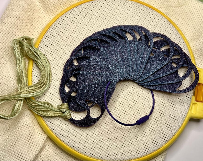 May include: A dark blue, fan-shaped needlework thread organizer with cut-out designs, held together by a purple cord and clasp. The organizer rests on a yellow embroidery hoop with cream-colored fabric and green thread.