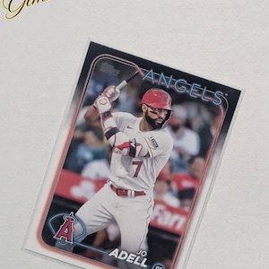 Peut inclure: Carte de baseball représentant Jo Adell des Angels de Los Angeles. La carte montre Adell en uniforme blanc avec des touches de rouge, en train de frapper avec une batte. Le logo des Angels et les détails du joueur sont visibles.