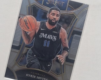 May include: A basketball trading card featuring Kyrie Irving, a basketball player for the Dallas Mavericks. The card is silver and blue with gold accents. The card features a photo of Irving dribbling a basketball. The card text reads "Select" and "Mavs 11".