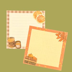 May include: Two square memo pads with autumn-themed designs. One pad features a brown gingham border, lined paper, orange citrus slices, and two lit candles. The other pad has an orange border with small stars, grid paper, a leaf, and books.