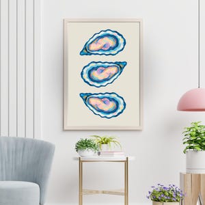 May include: A framed art print showing three oyster shell illustrations. The shells are painted with blue, yellow, and pink colours on a cream background. The artwork is presented in a light-coloured wooden frame.