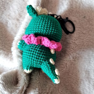 May include: Green crocheted dinosaur keychain with a pink ruffle collar and white spots. The keychain has a black metal ring.