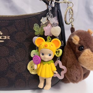 Puede incluir: Un llavero con una figura amarilla con tem&aacute;tica de rat&oacute;n que sostiene una piruleta, con una colecci&oacute;n de amuletos que incluyen mariposas, estrellas y corazones. Un llavero de oso de peluche marr&oacute;n cuelga cerca.