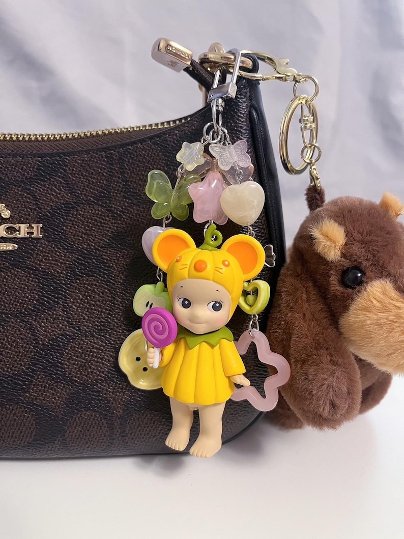 Puede incluir: Un llavero con una figura amarilla con tem&aacute;tica de rat&oacute;n que sostiene una piruleta, con una colecci&oacute;n de amuletos que incluyen mariposas, estrellas y corazones. Un llavero de oso de peluche marr&oacute;n cuelga cerca.