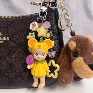 Puede incluir: Un llavero decorativo con una figura amarilla con disfraz de rat&oacute;n sosteniendo una piruleta, un oso de peluche marr&oacute;n y varios abalorios. El llavero est&aacute; sujeto a un bolso marr&oacute;n con herrajes dorados.